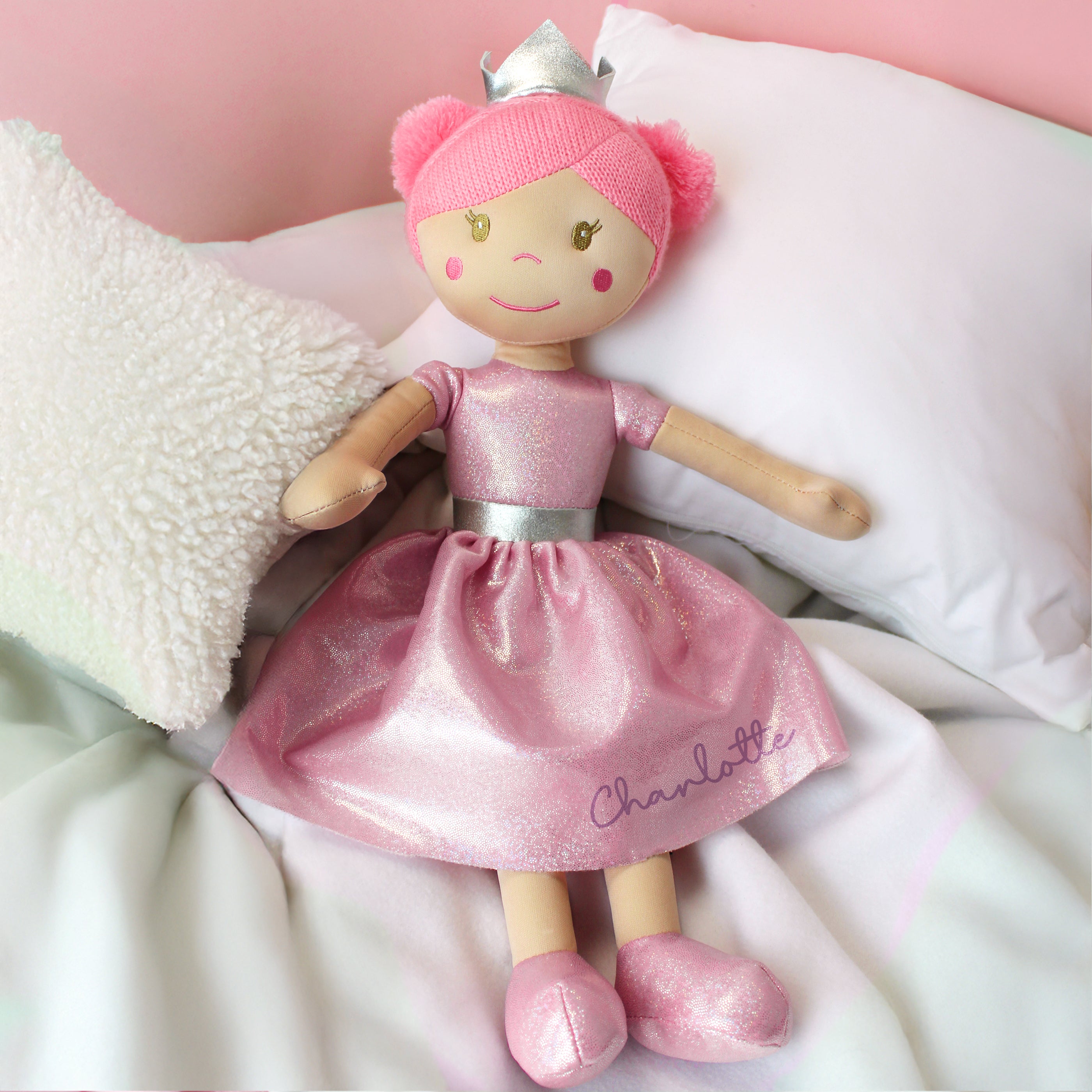 Doll in a pink dress with 'Charlotte' on it, wearing a tiara, on a soft surface.
