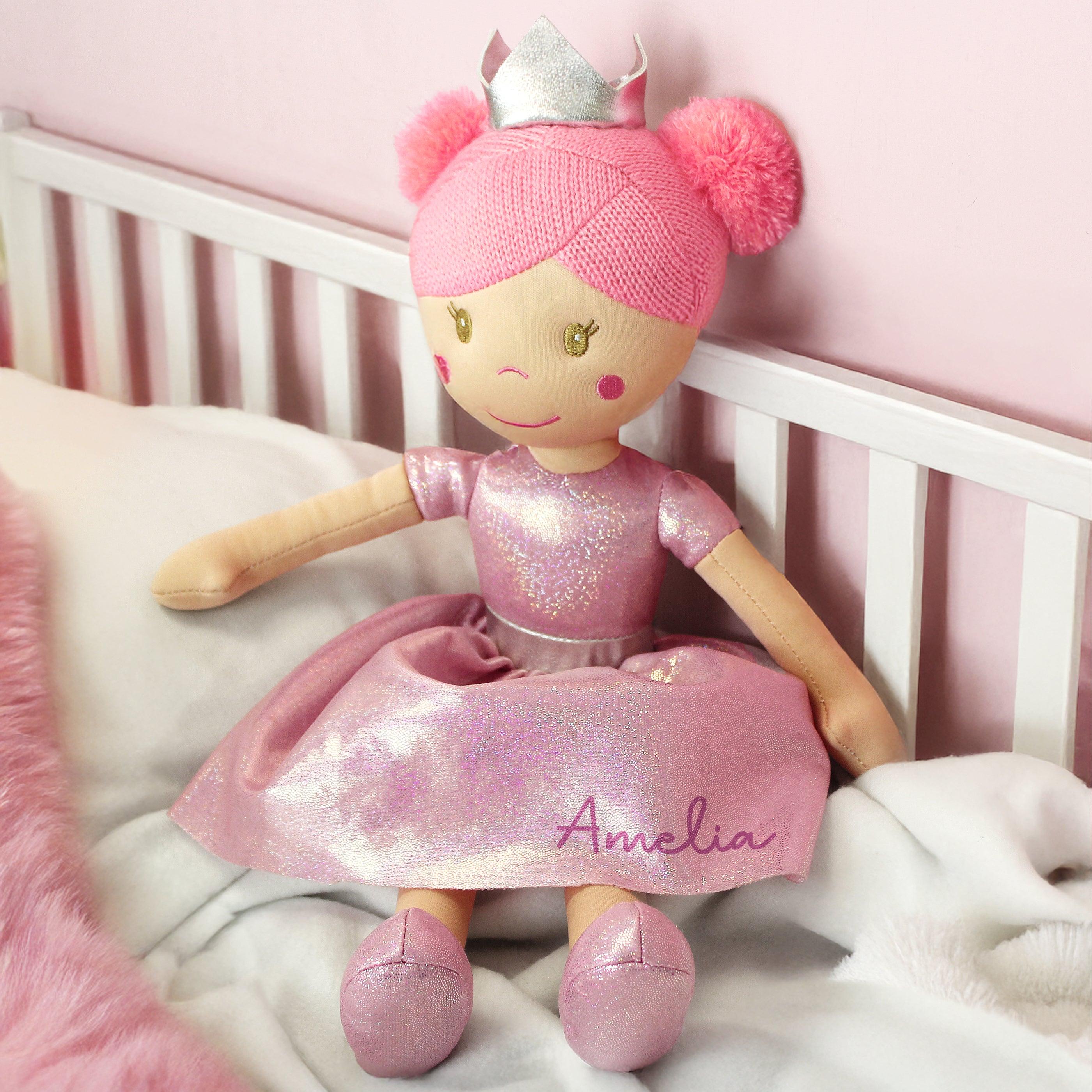 Doll named Amelia in a pink outfit with a tiara, sitting on a white crib with a pink background.