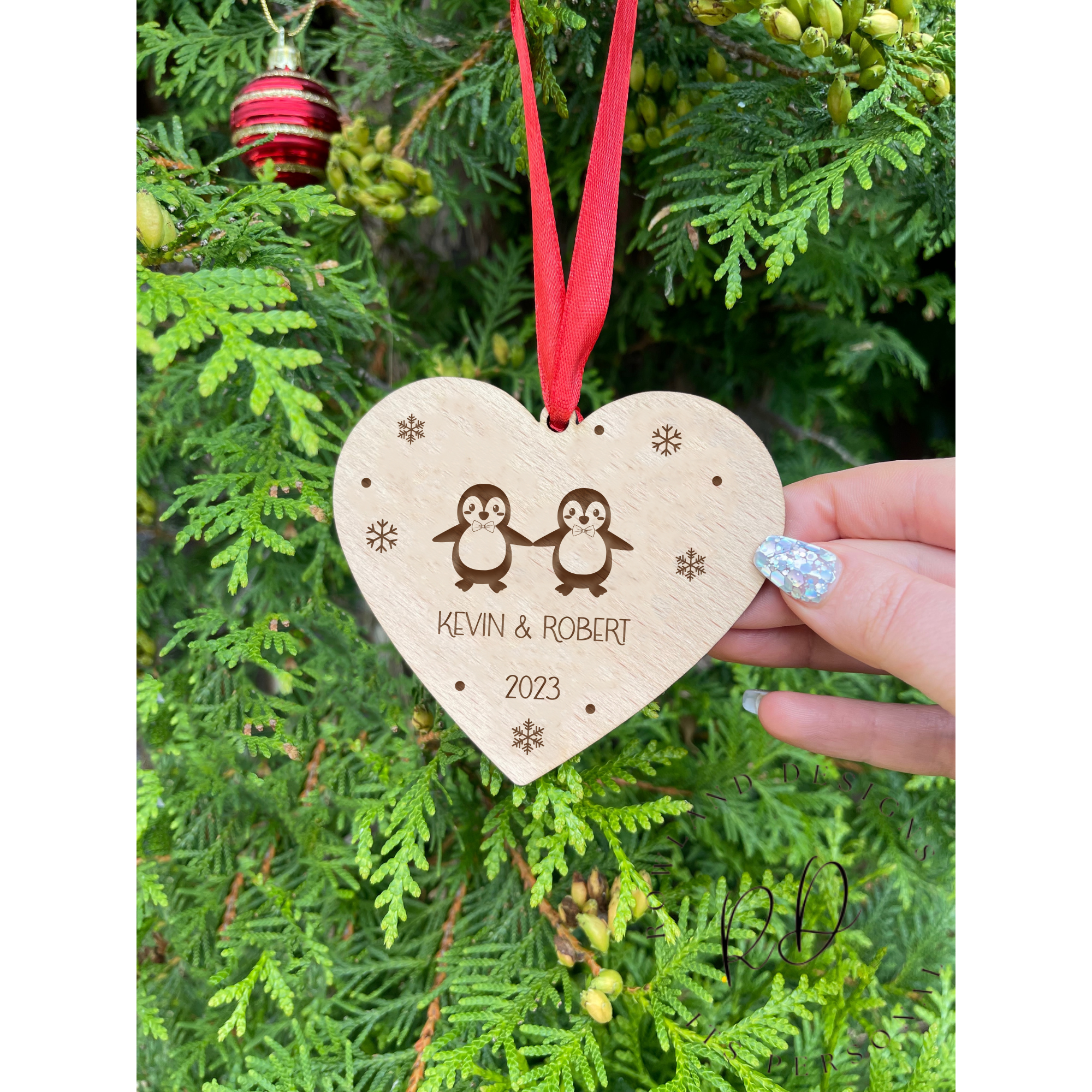Close-up of a personalised wooden heart-shaped ornament for same-sex male couples, featuring two penguins with bow ties, custom names, and a year engraving.