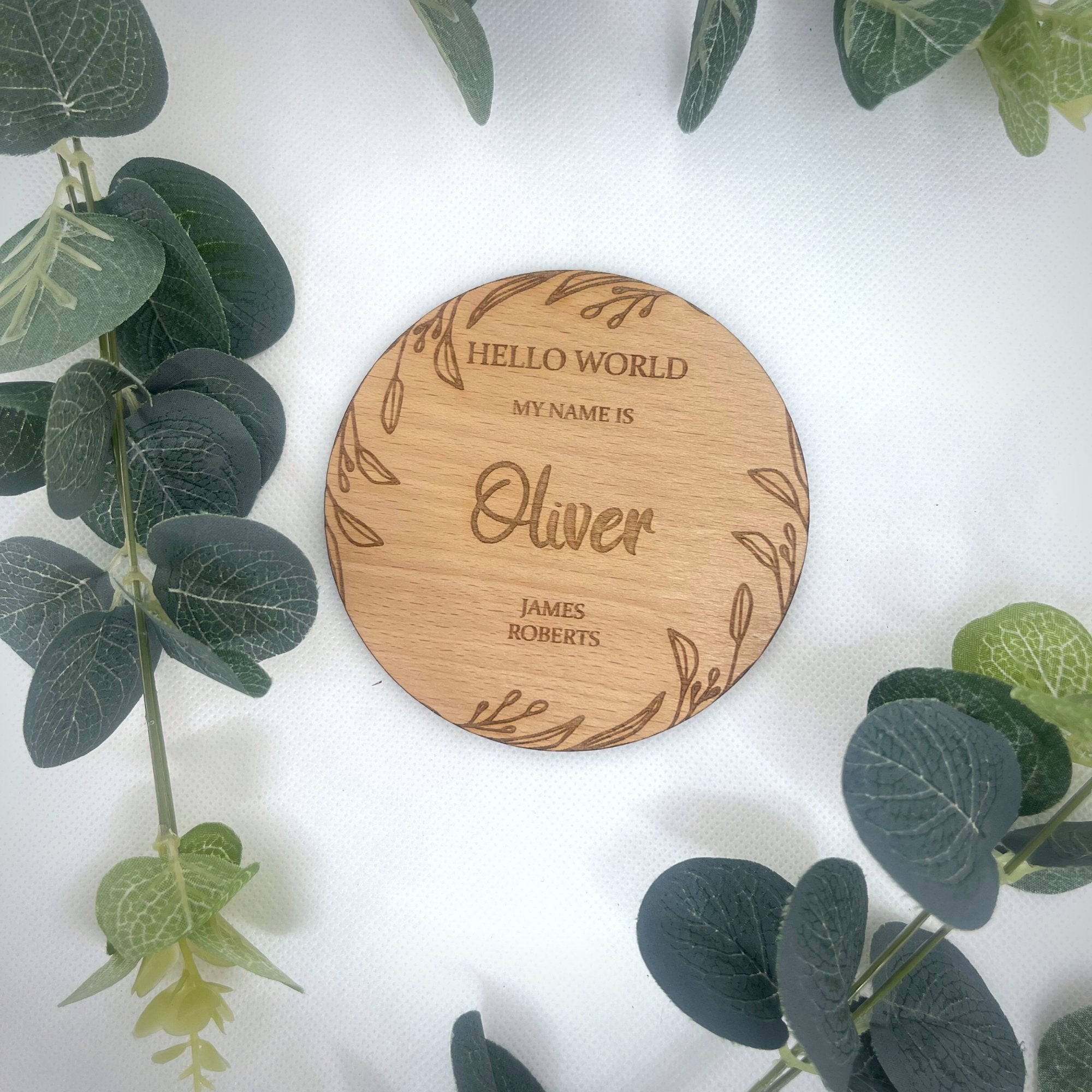 A personalised round wooden plaque with natural leaf design, engraved with the text 'Hello World' and a name, on a white background with green leaves in the background.