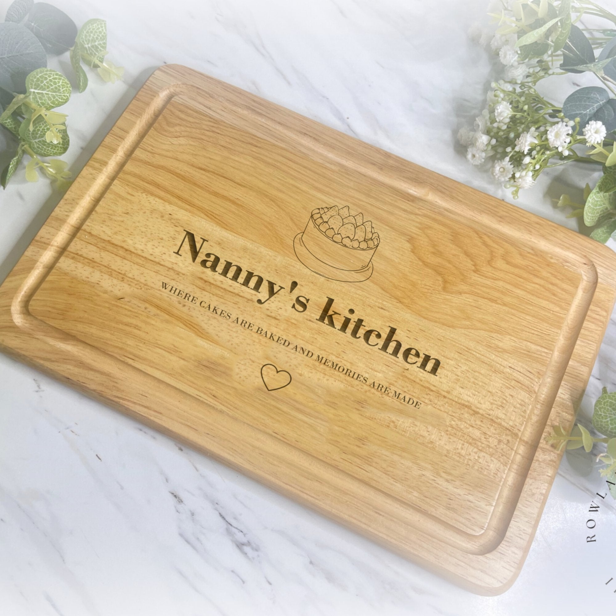 A personalised wooden chopping board with the inscription 'Nanny's kitchen' along with a cake icon and a heart symbol engraved on it.