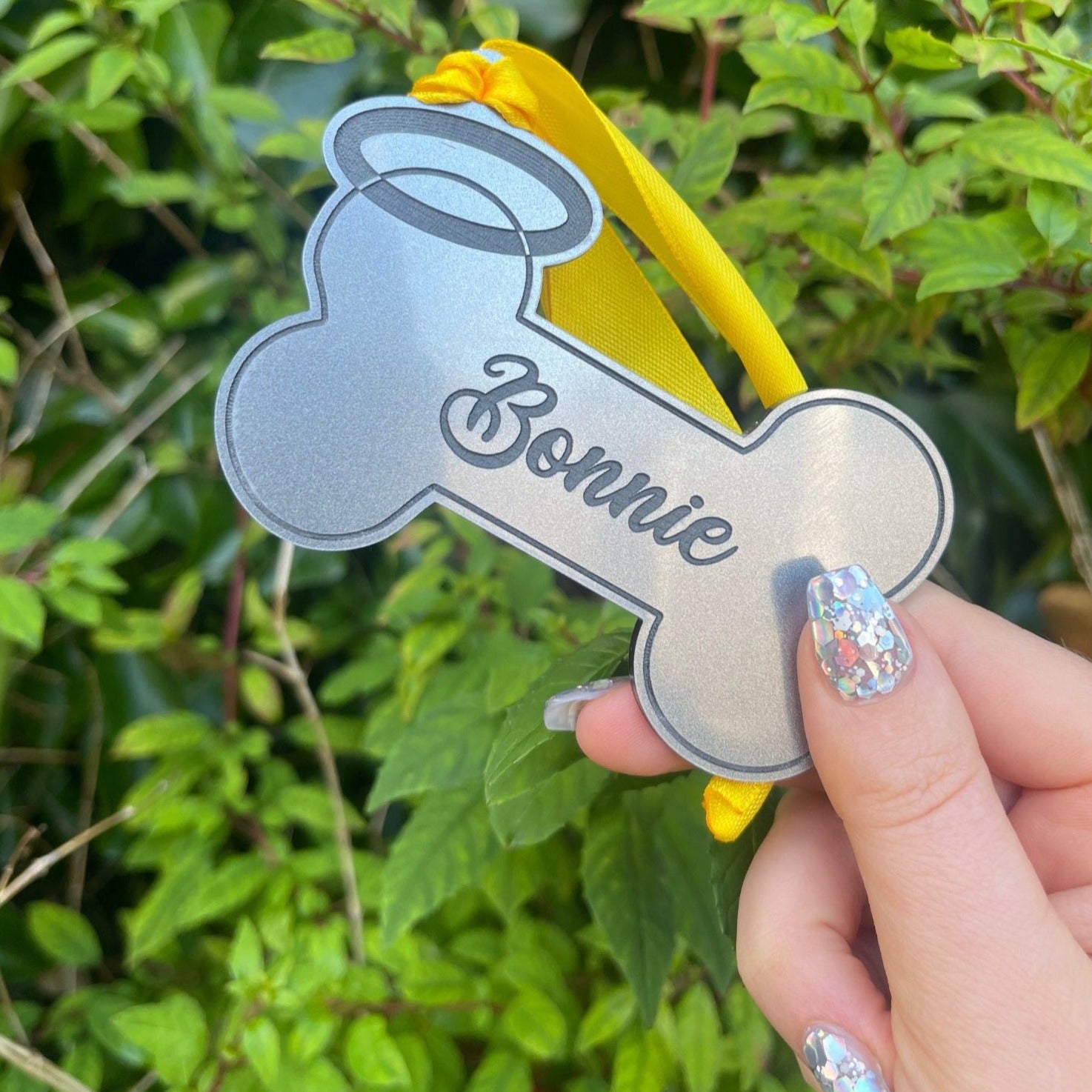 A personalised dog memorial gift in the form of a hanging bone ornament with a yellow ribbon and black engraving of a dog's bone, with the name 'Bonnie'.