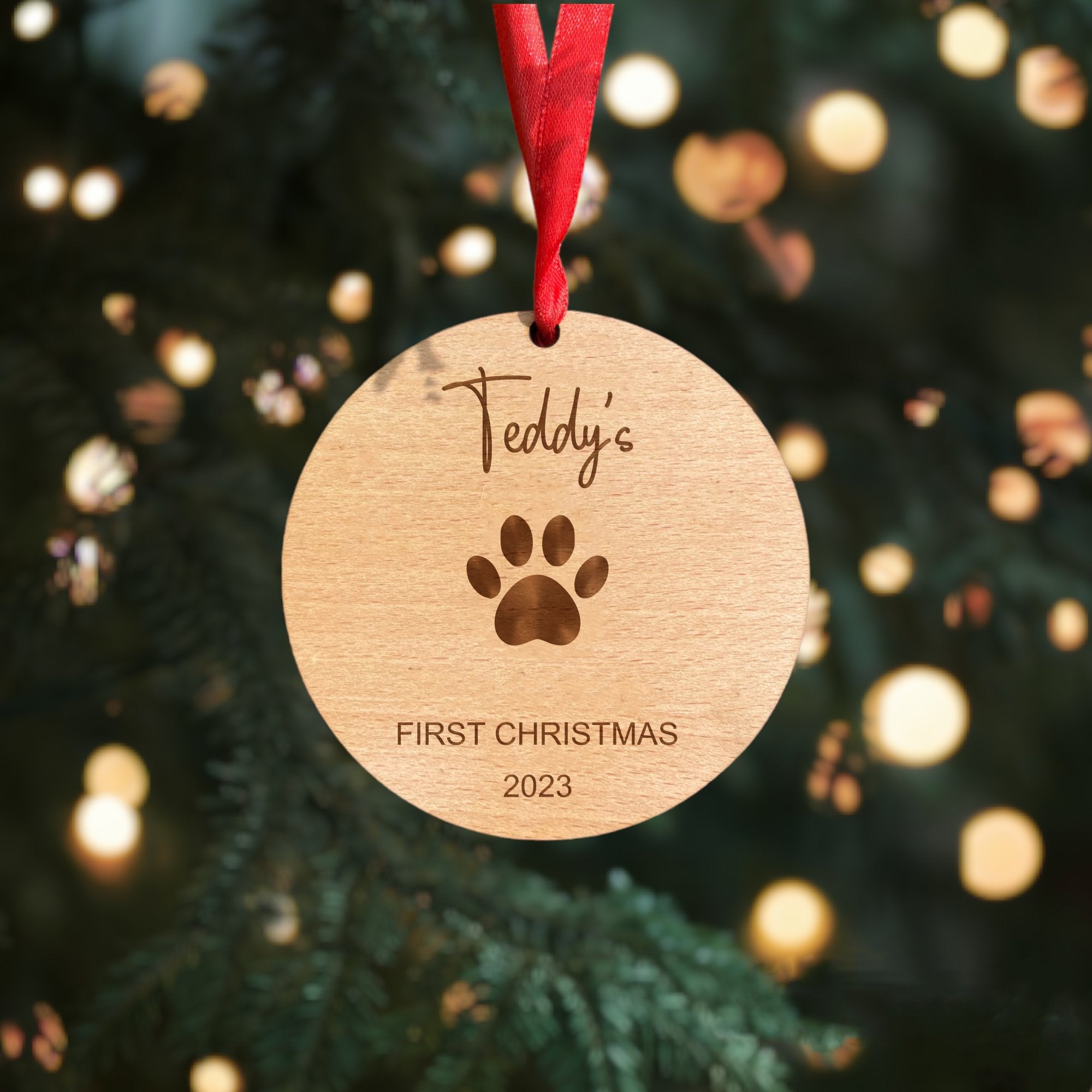 Personalised wooden round Christmas bauble with a paw print and 'First Christmas' engraving, with a red ribbon.