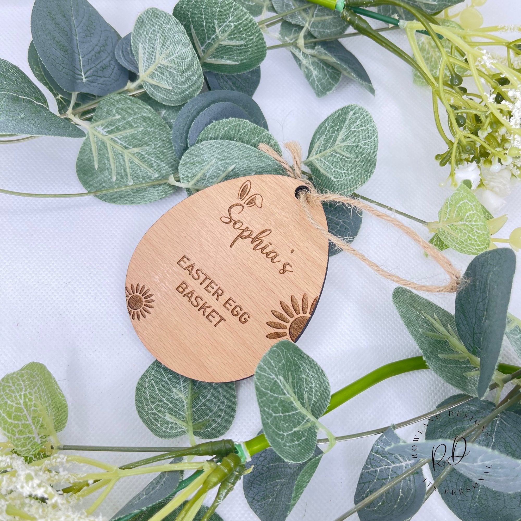 A personalised wooden Easter egg-shaped tag with the text 'Sophia's Easter Egg Basket' engraved on it, placed on a bed of green leaves.