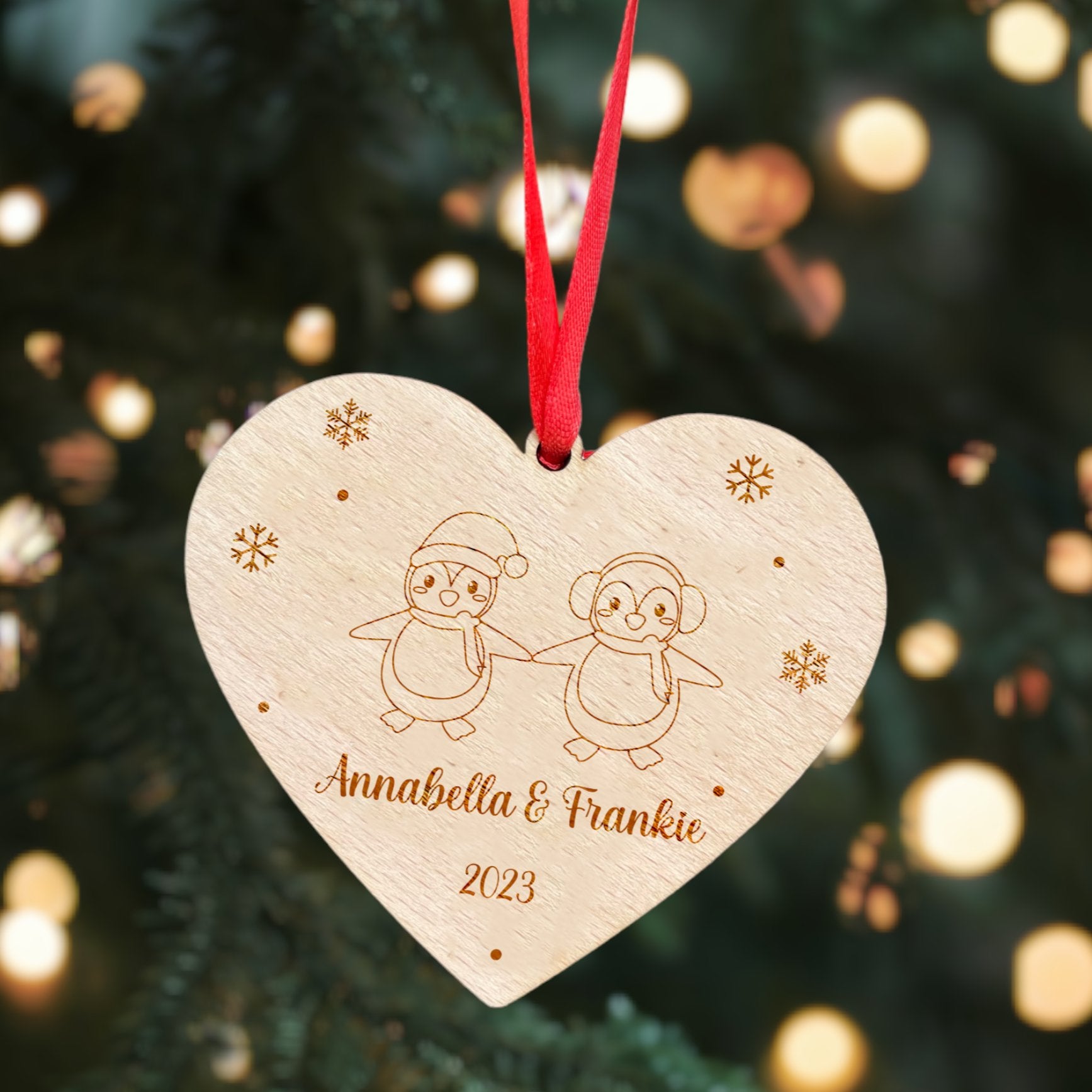 A heart-shaped wooden Christmas ornament with engraved penguins and snowflakes, hanging from a red ribbon.