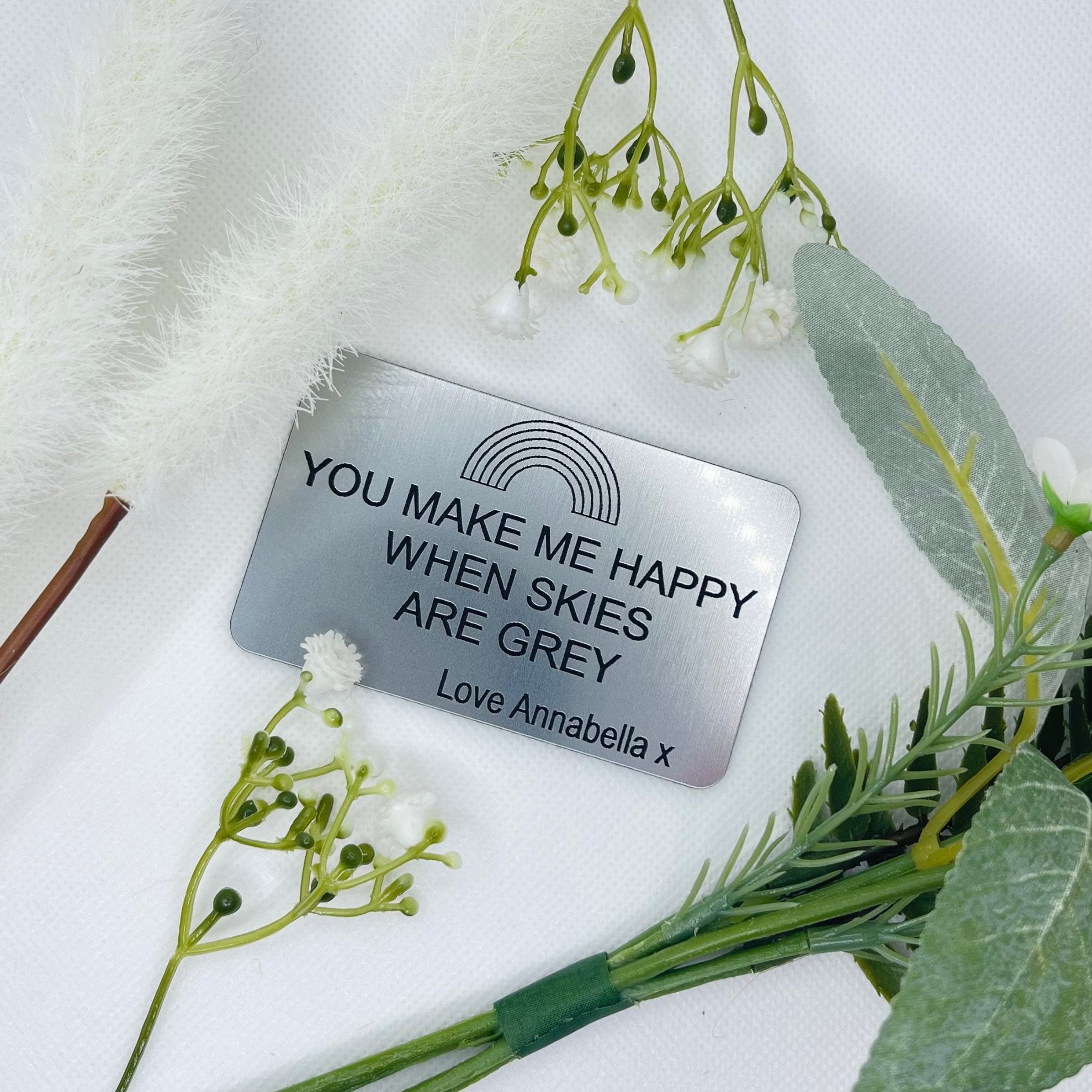 A personalized clear acrylic wallet card insert with the text 'YOU MAKE ME HAPPY WHEN SKIES ARE GREY' engraved on it, placed on a surface with decorative flowers around it.