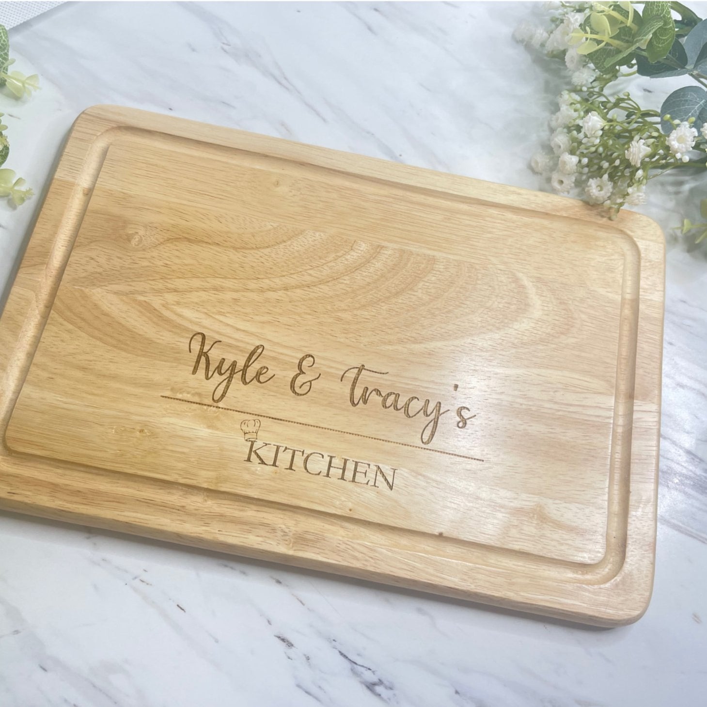 A personalised wooden chopping board with an engraved chef hat symbol and custom text 'Kyle & Tracy's Kitchen'.
