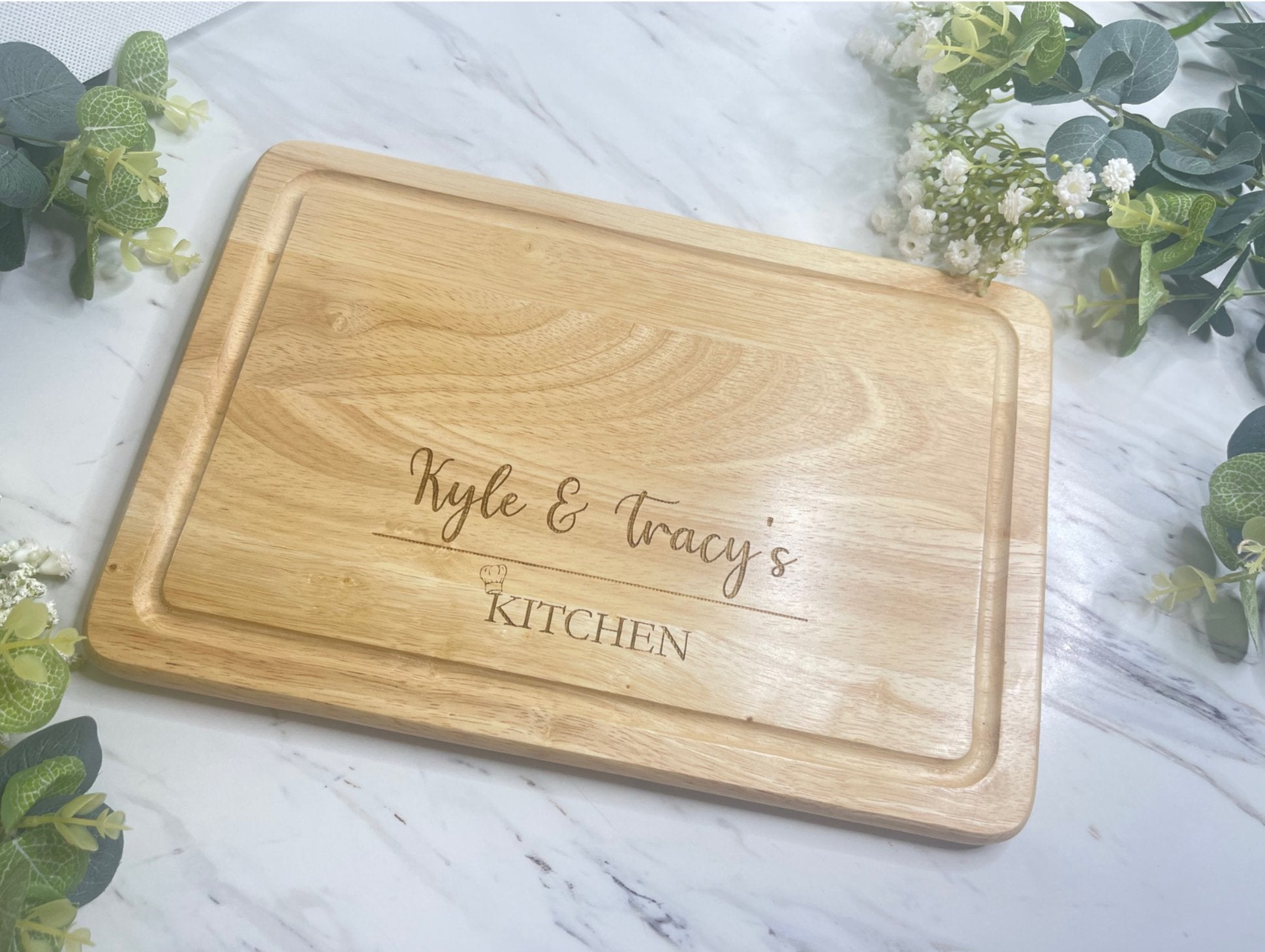 A personalised wooden chopping board with an engraved chef hat symbol and custom text 'Kyle & Tracy's Kitchen'.