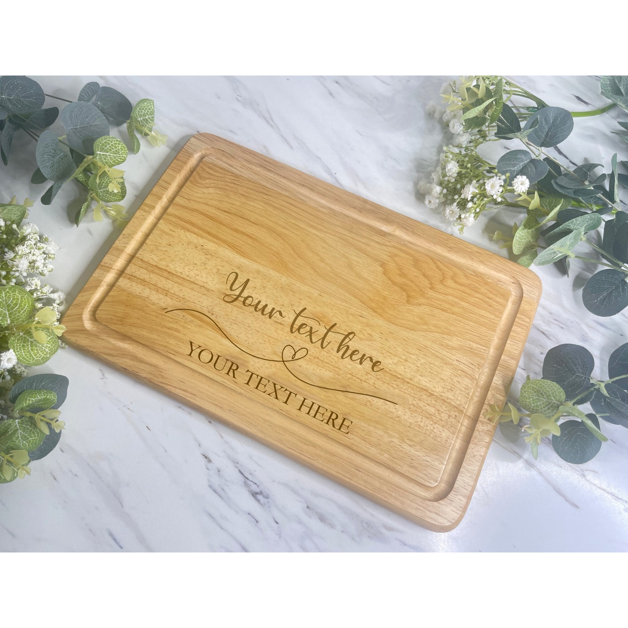 A personalised wooden chopping board with heart designs, engraved with text 'Your Text Here' on a marble surface with green foliage in the background.
