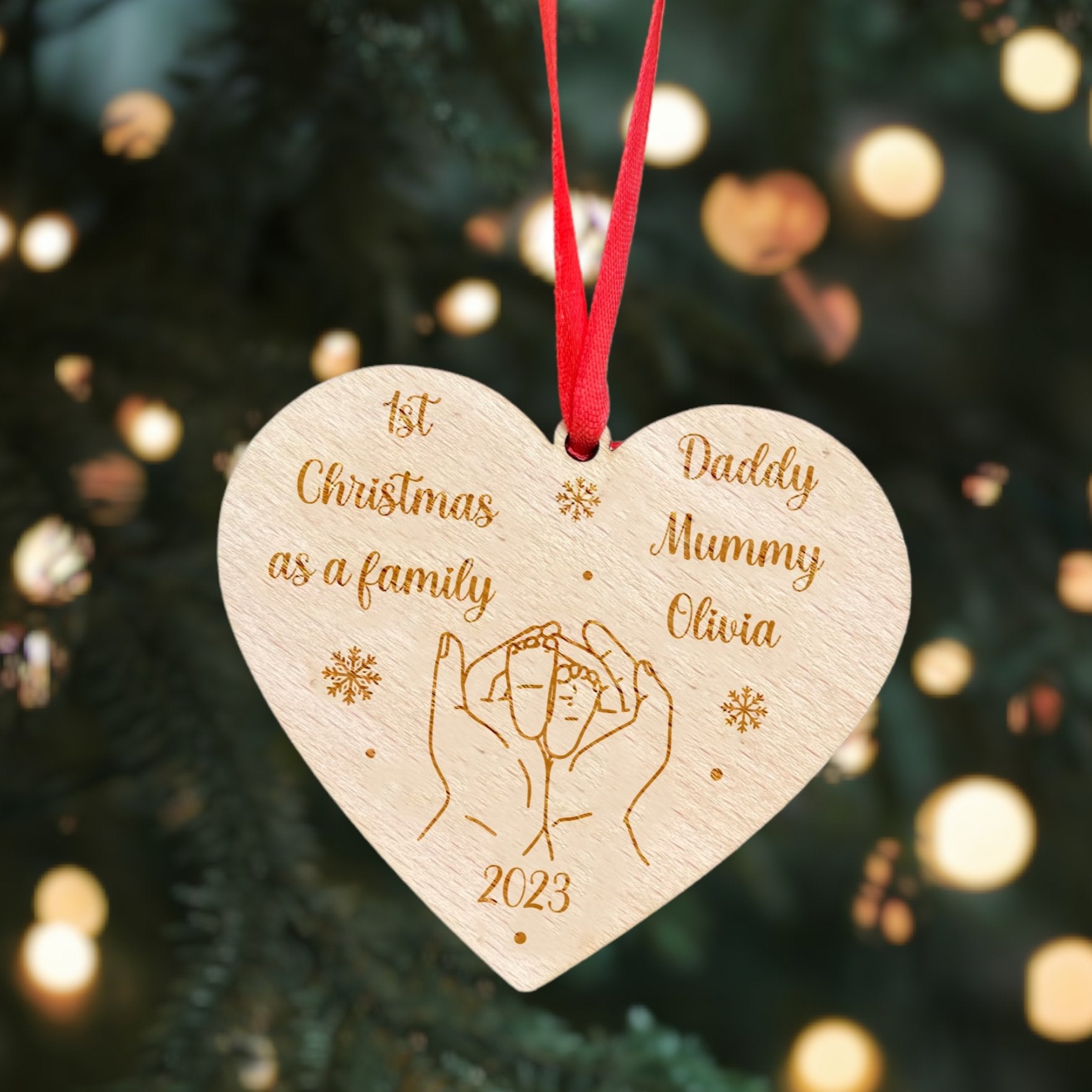 A heart-shaped wooden Christmas ornament with '1st Christmas as a Family' engraved on it, along with a couple's hands intertwined and the year 2023 at the bottom, adorned with a red ribbon.