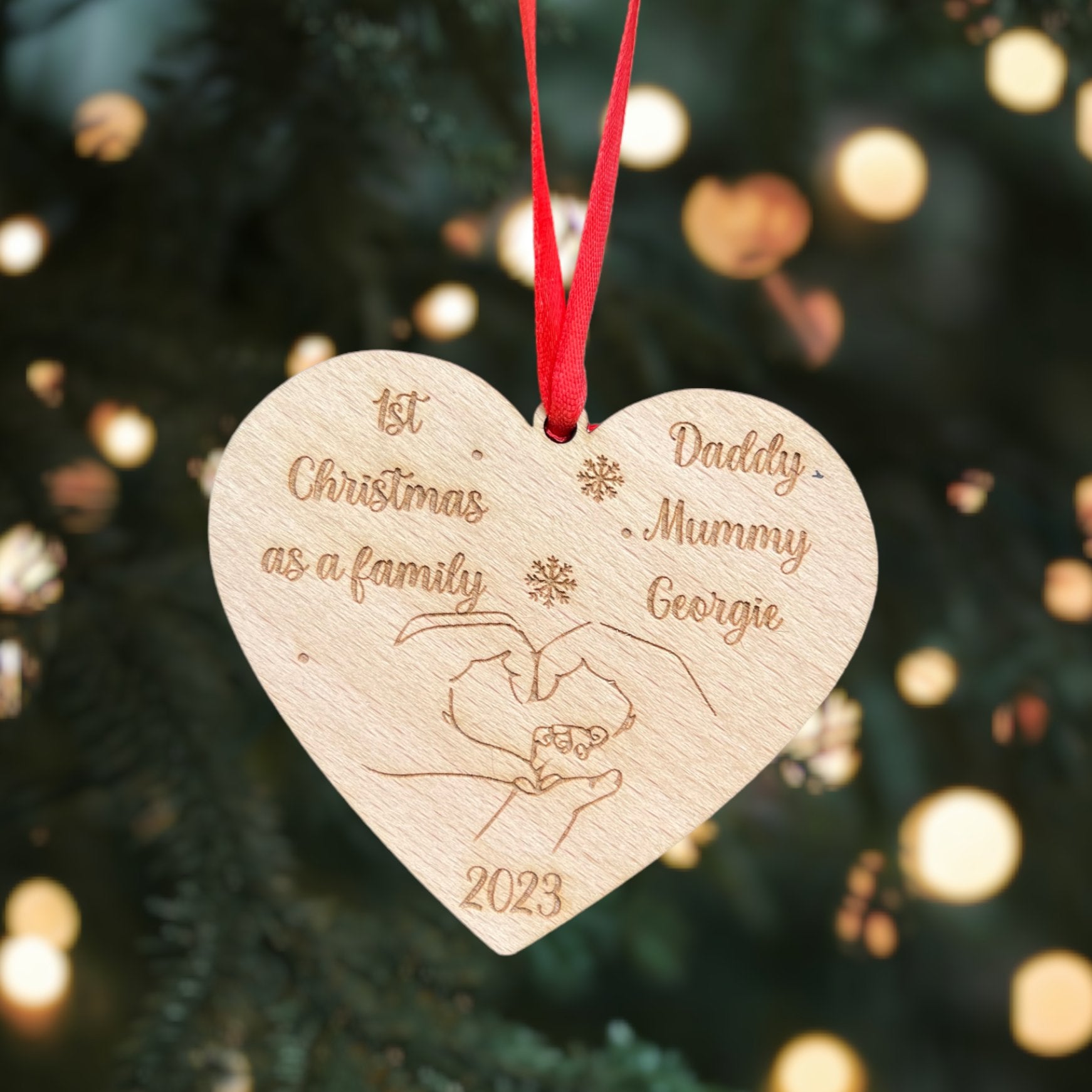 A personalised wooden heart-shaped Christmas bauble with engraved text '1st Christmas as a Family' and spaces for personal names. It is hanging on a Christmas tree with a red ribbon.