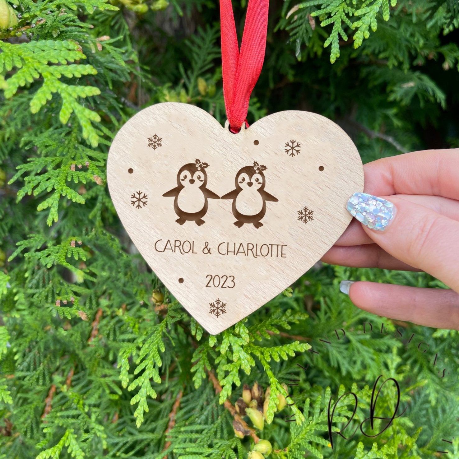 Close-up of a personalised wooden heart-shaped ornament for same-sex couples, featuring two penguins with bows, custom names, and a year engraving.
