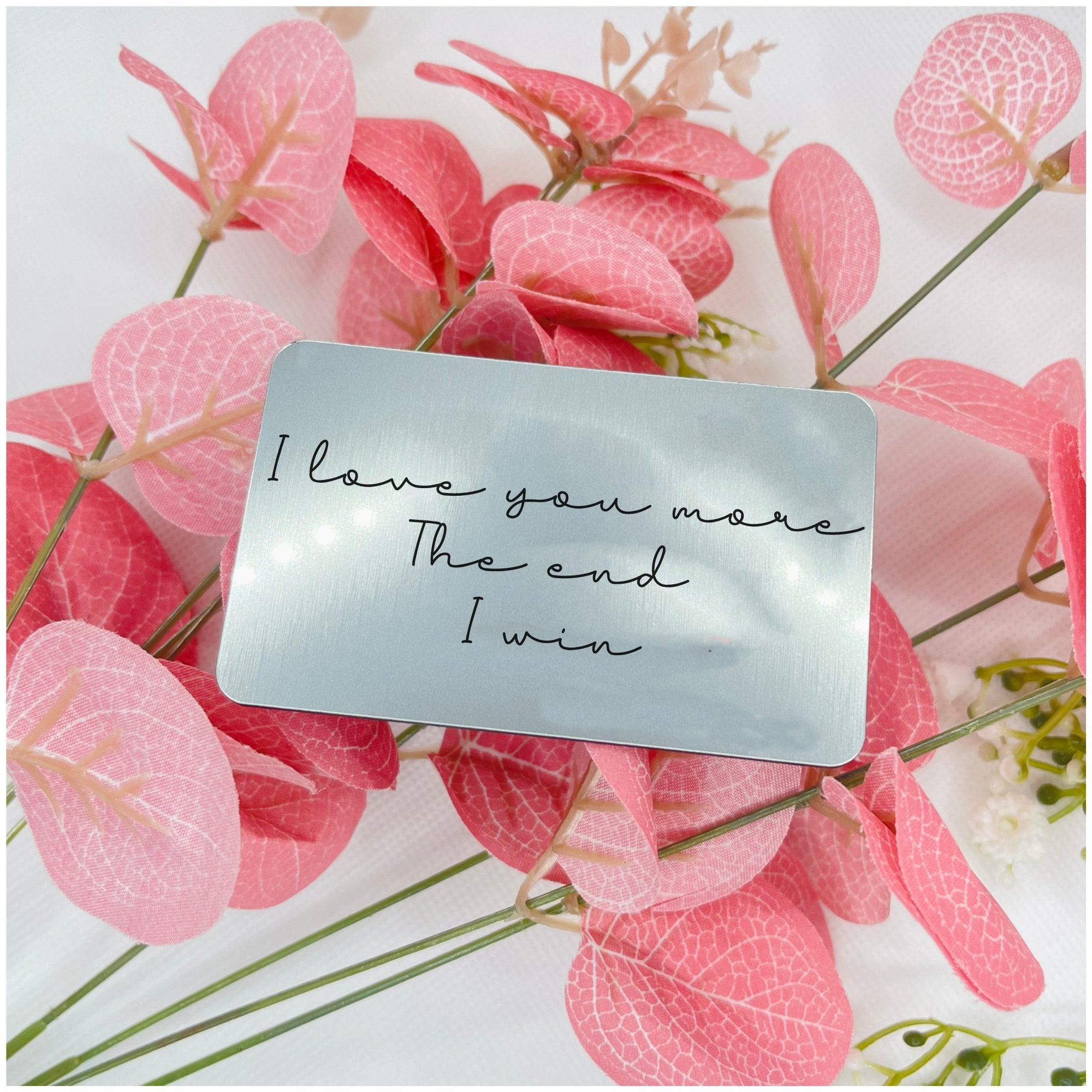 A silver wallet card with the engraving 'I love you more The end I win' placed on a background of pink flowers.