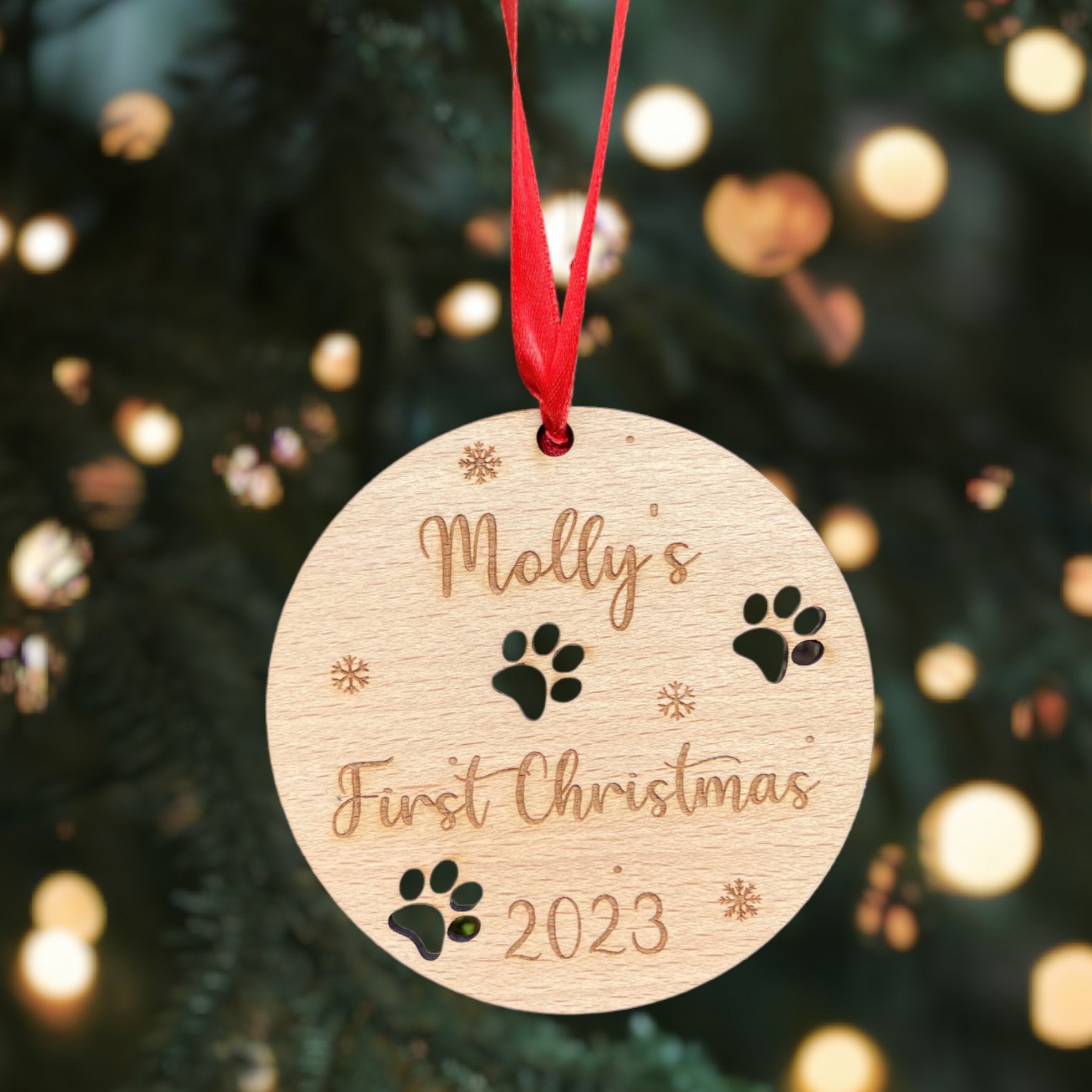 A personalised wooden round Christmas bauble with a paw print and 'First Christmas ' engraving, hanging from a red ribbon.