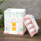 A soft blush pink ceramic oil burner and wax warmer with a cozy house design. Features a removable top and a tealight holder to release calming scents into your home.