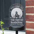 Halloween-themed welcome sign with a haunted house design on a black stand, placed against a door and brick wall.