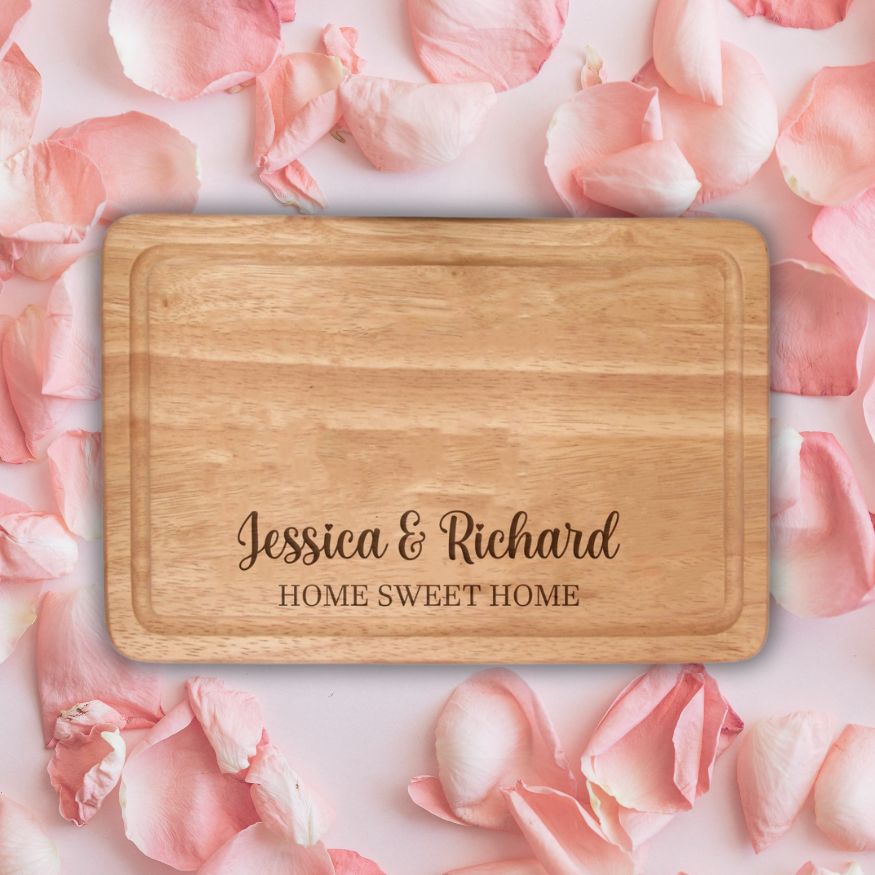 Wooden cutting board with engraved text 'Jessica & Richard HOME SWEET HOME' on pink rose petals.