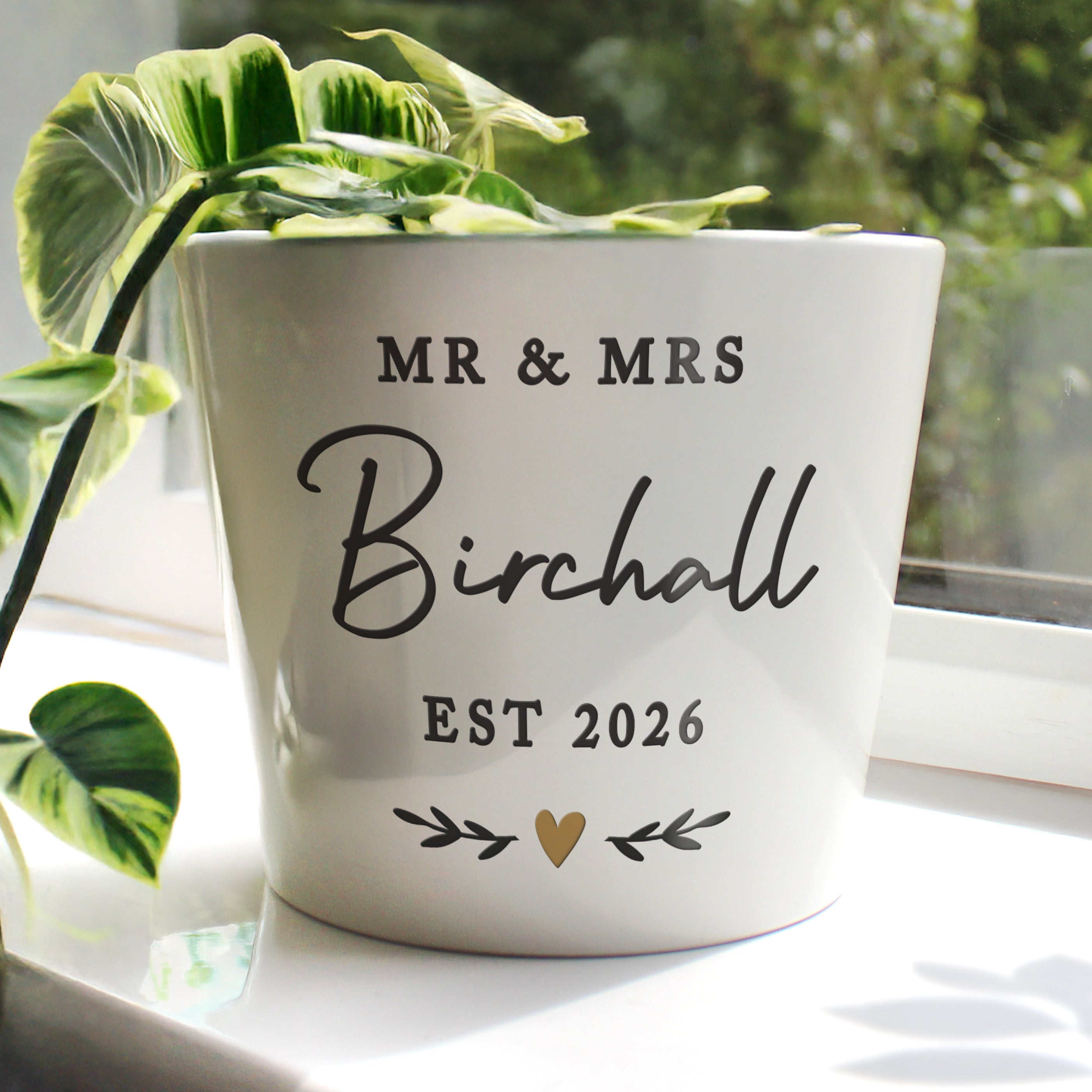 White planter with 'MR & MRS Birchall EST 2026' text on a light surface with green leaves in the background.