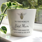 White pot with bee design and message to 'The World's Best Mum' on a light surface with green leaves in the background.