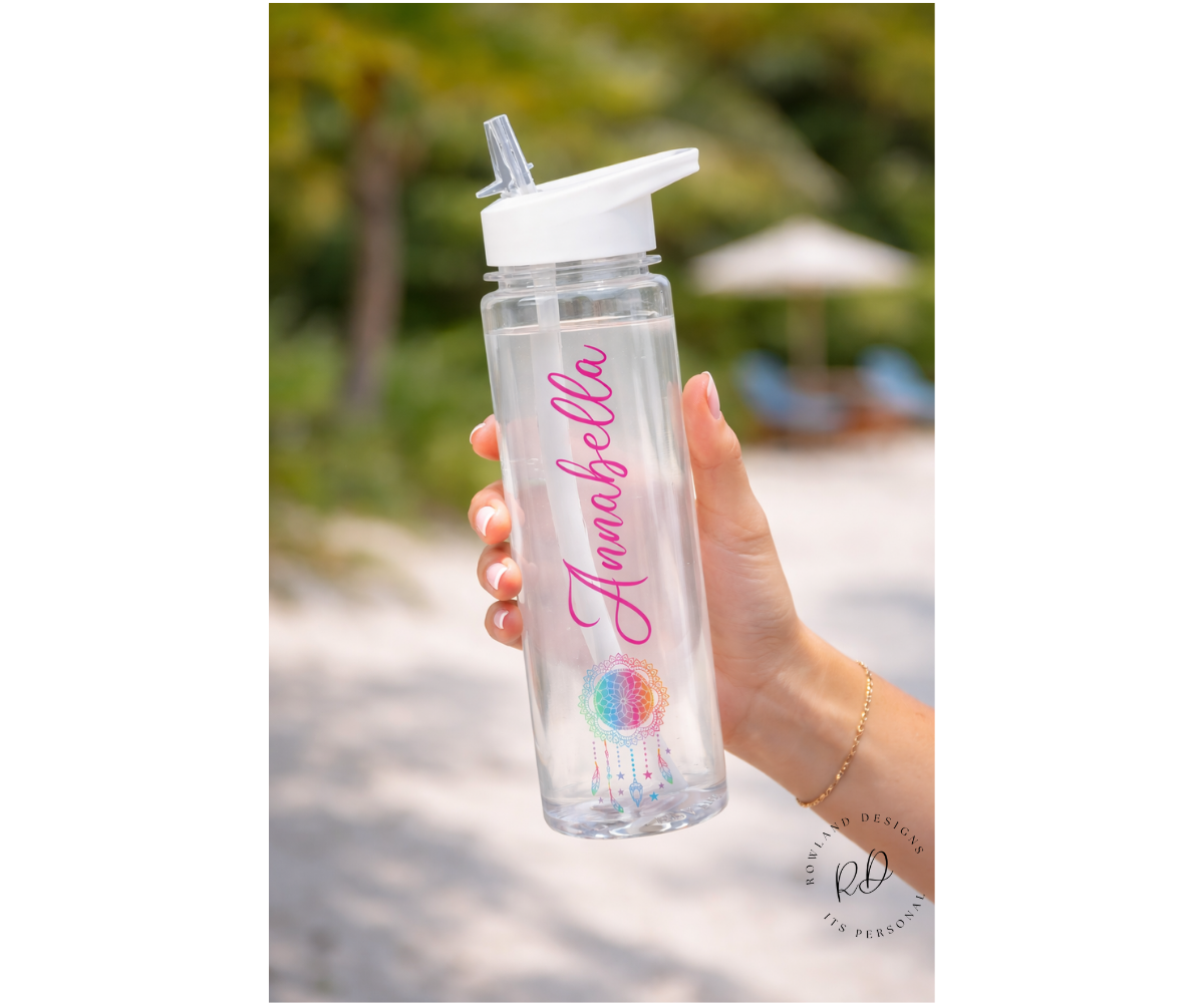 Clear water bottle with pink name and rainbow dream catcher design held by a hand outdoors.