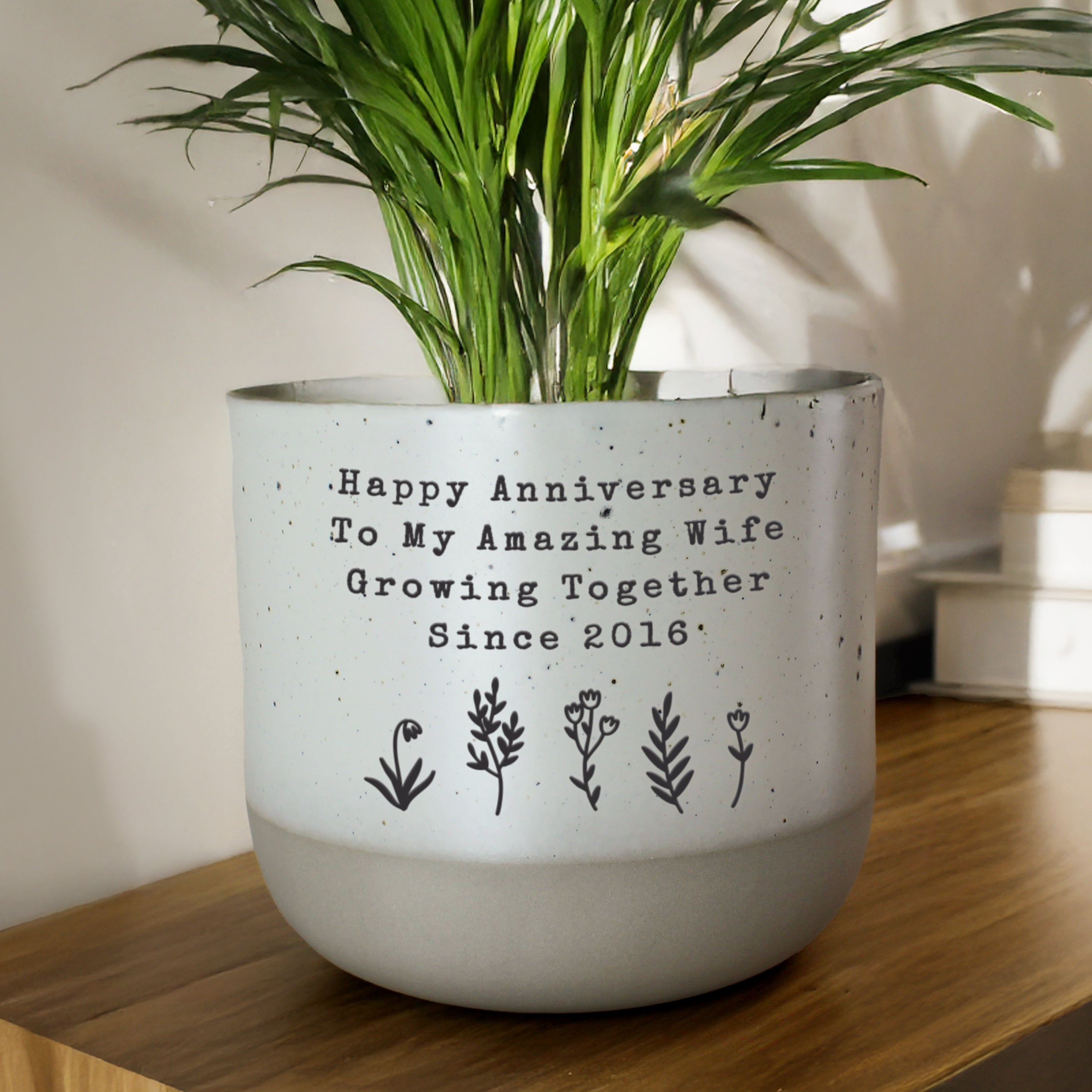 White planter with green plant and customised anniversary message on a wooden surface.