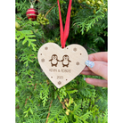 Close-up of a personalised wooden heart-shaped ornament for same-sex male couples, featuring two penguins with bow ties, custom names, and a year engraving.