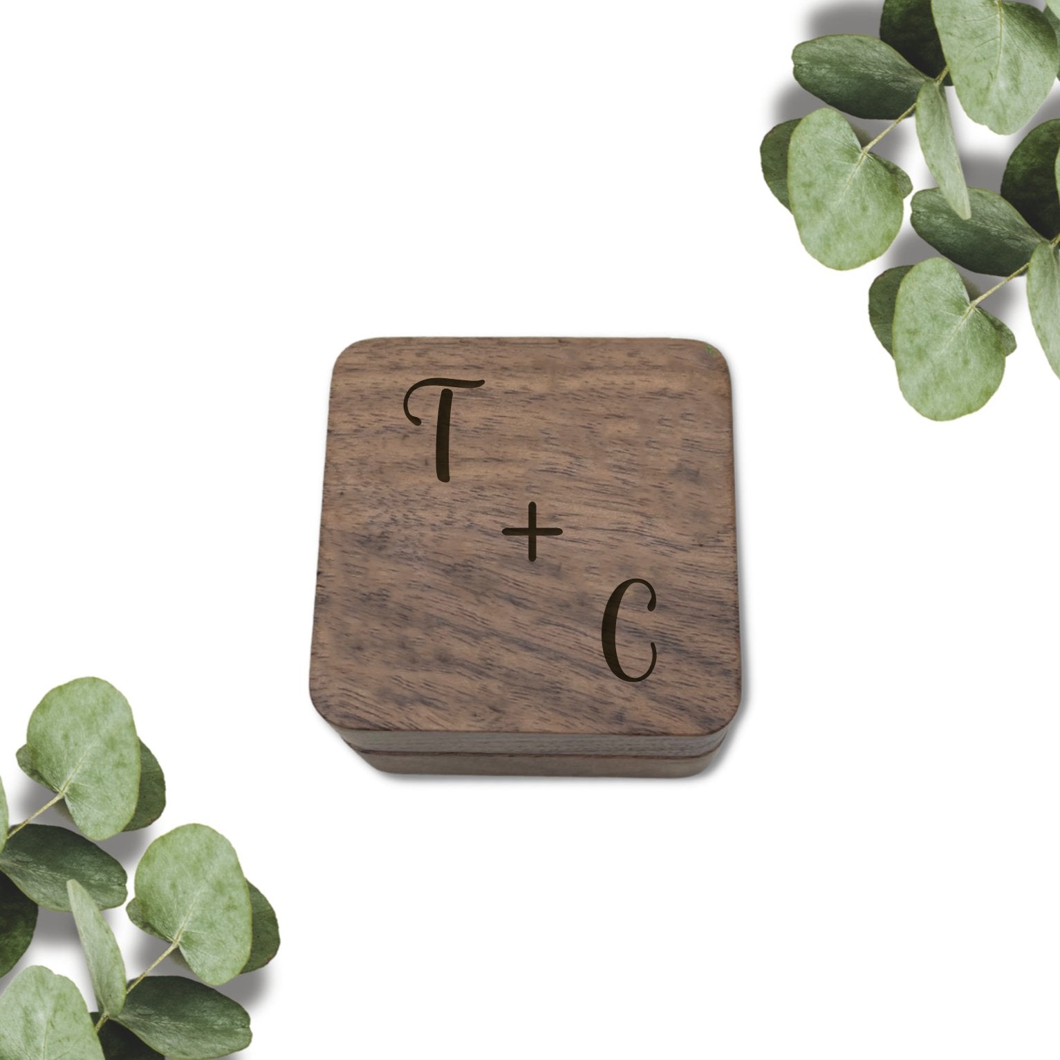 Personalised double slot wooden ring box with the initials 'T + C' engraved on top, placed on a white background with green leaves around it.